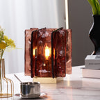 A lit Clérachic table lamp with textured amber-red glass panels glows on a marble tabletop, beside a steel cup and a vase of yellow flowers.