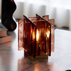 A Clérachic table lamp with tall amber-red glass panels glows warm on a white marble surface, with a dark vase and books behind it.