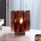 Close up of a lit Clérachic table lamp, with a candle-style bulb glowing through its amber-red glass panels, in front of a sheer curtain.