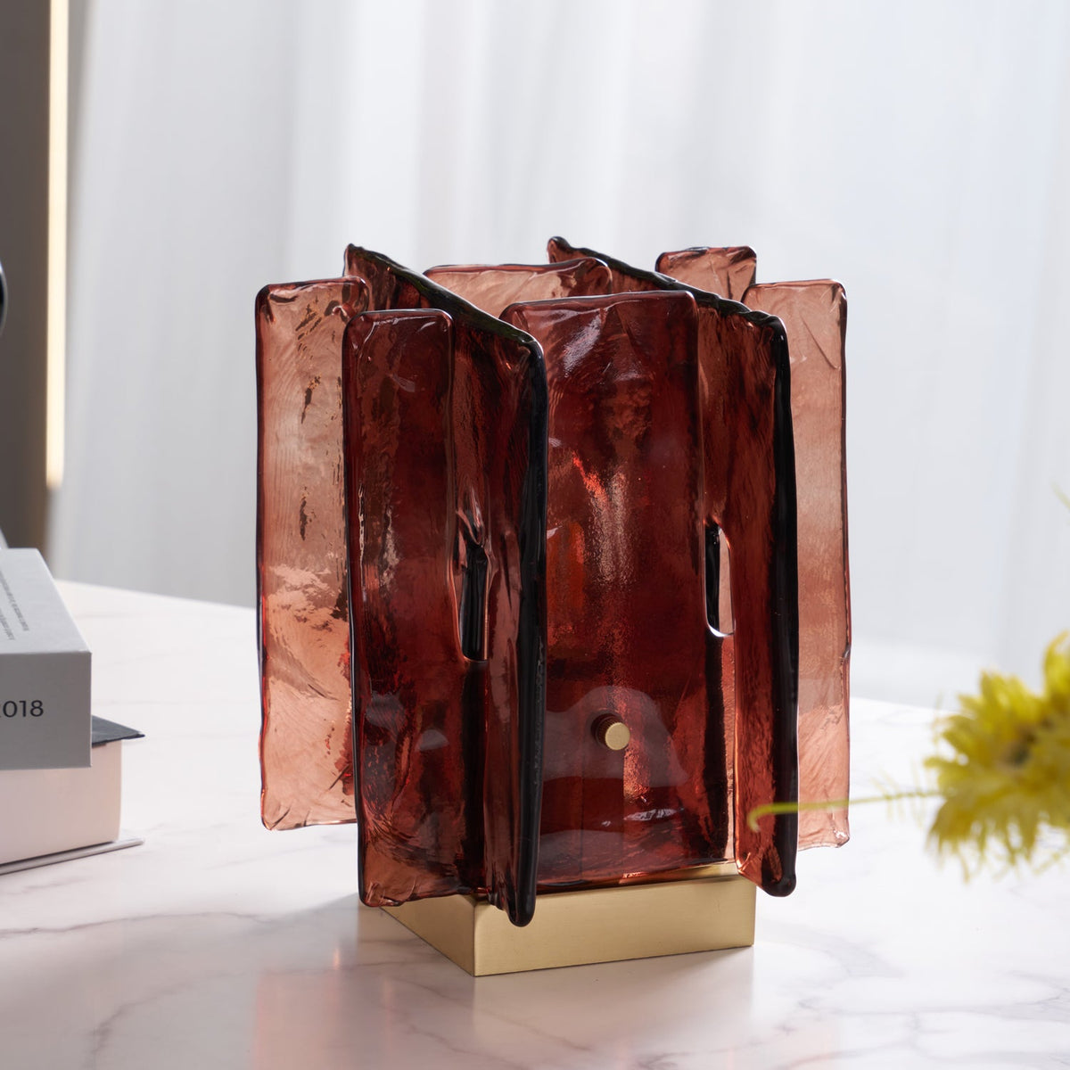 Unlit, this Clérachic table lamp shows its translucent amber-red glass panels and brass base on a marble table, with yellow flowers beside it.