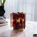A Clérachic table lamp with glowing amber-red glass panels and a brass base sits on a marble table, with a black vase and books to the left.