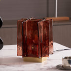 Unlit, a Clérachic table lamp with deep amber-red glass panels and a brass base stands on marble, with a small steel cup beside it.