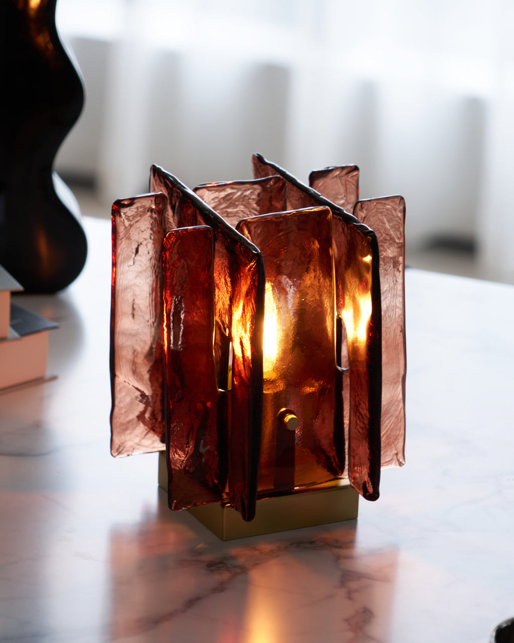 A lit Clérachic table lamp glows warm through its overlapping amber-red glass panels, on a marble surface in front of a curtain.