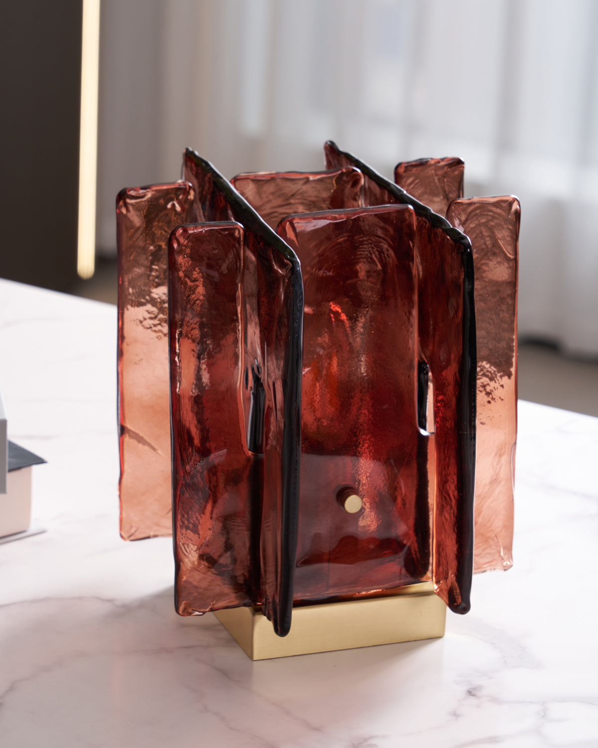 A Clérachic table lamp with amber-red glass panels, seen at an angle on a white marble table, with a small brass pin on the front panel.