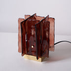 An unlit Clérachic table lamp with layered amber-red glass panels and a brass base, with a black cord at the side, on a pale backdrop.