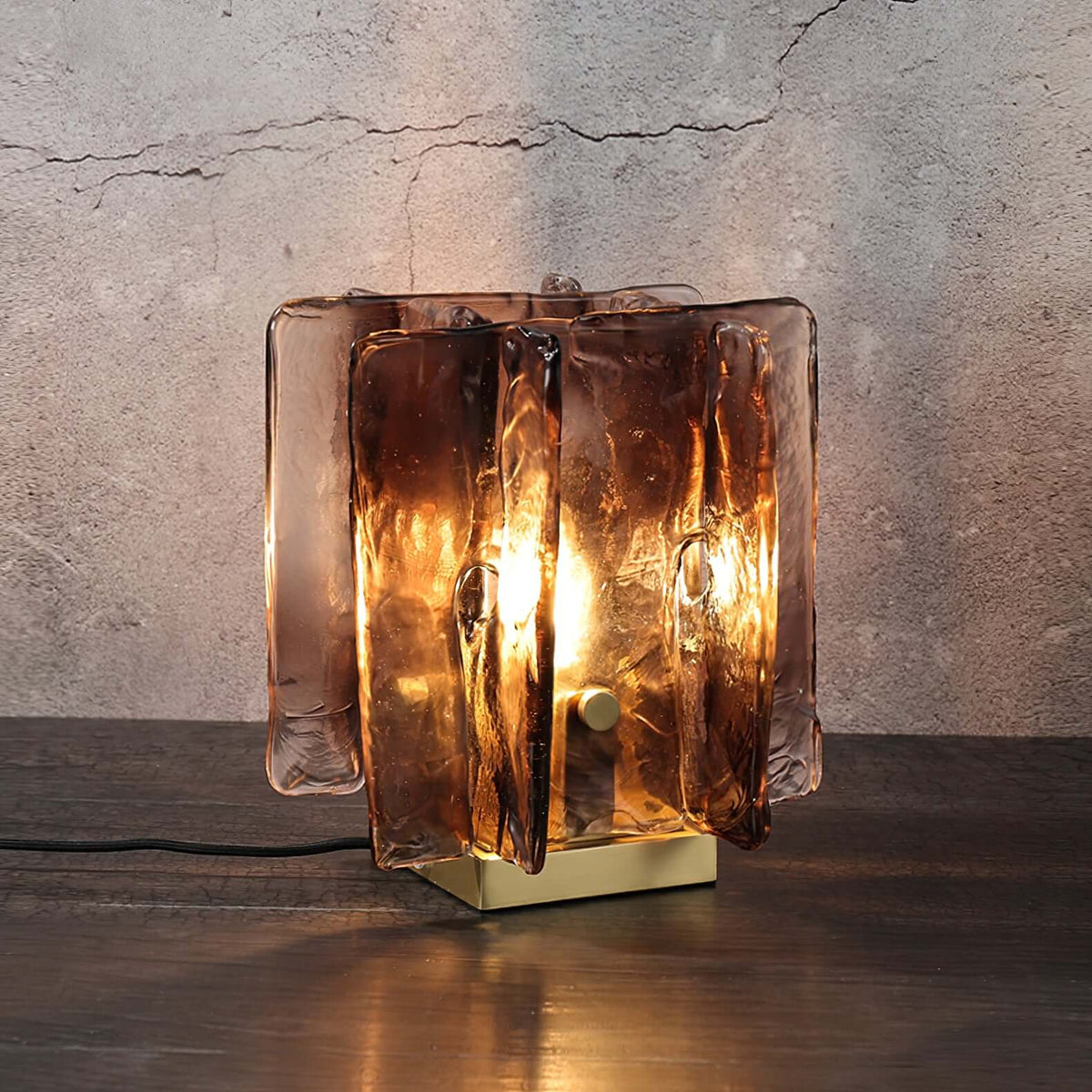 A lit Clérachic table lamp glows on a dark wooden shelf against a cracked grey plaster wall, its glass panels turning a smoky amber.
