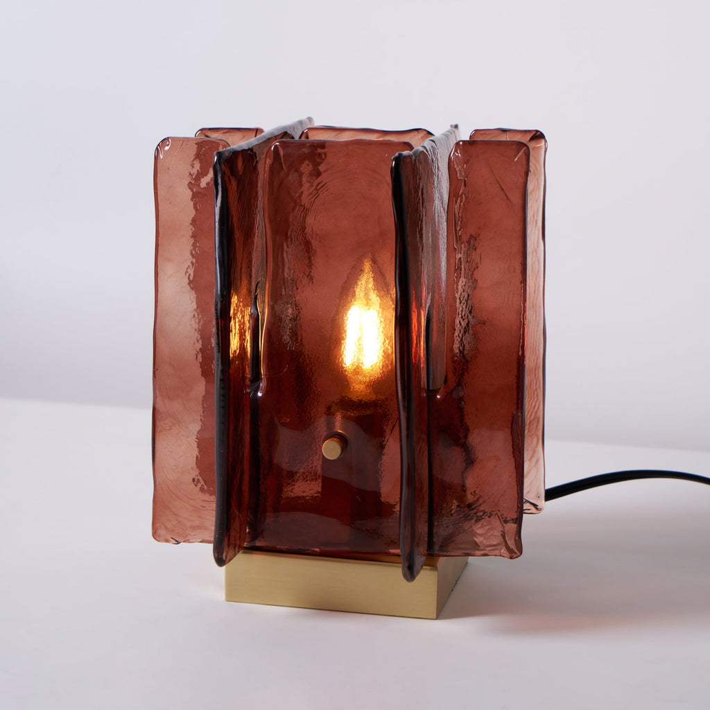 Straight-on view of a lit Clérachic table lamp, with a candle-style bulb glowing inside its amber-red glass panels and a black cord at the side.