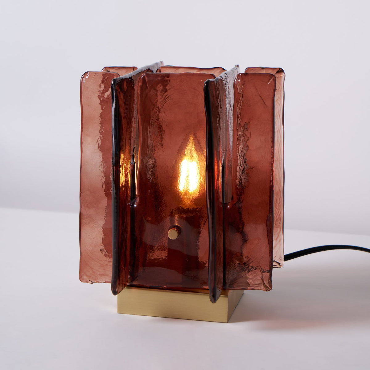 Straight-on view of a lit Clérachic table lamp, with a candle-style bulb glowing inside its amber-red glass panels and a black cord at the side.