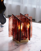A lit Clérachic table lamp with amber-red glass panels glows on a veined marble tabletop, with a dark vase and a curtain behind.