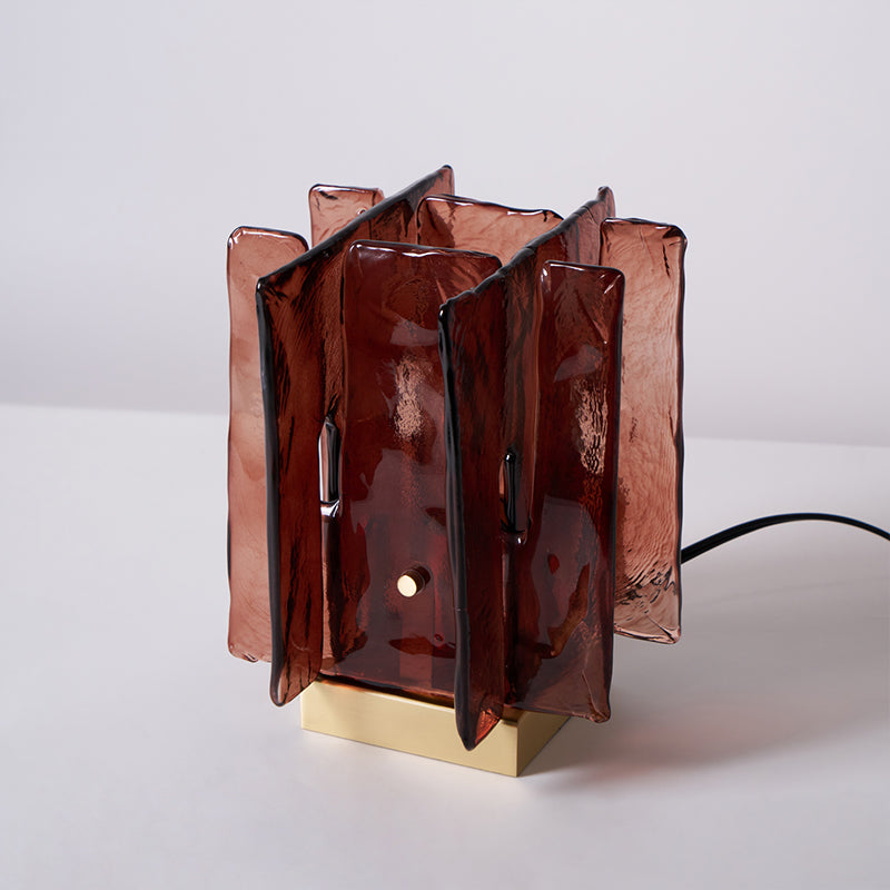 An unlit Clérachic table lamp with amber-red glass panels and a brass base stands on a white tabletop with its black cord trailing to the right.
