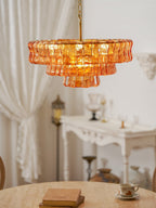 Soft-focus view of an amber Clérachic chandelier above a pink-beige round table, with white curtains and a console beyond.