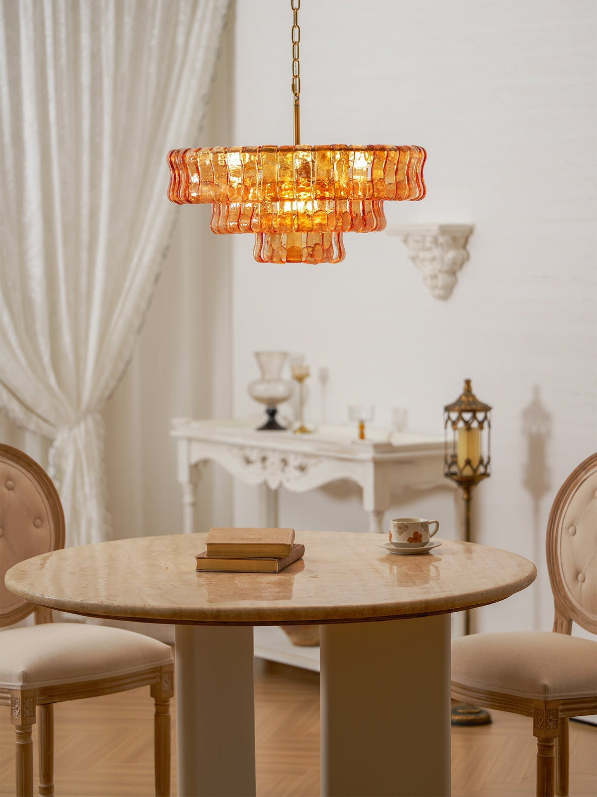 A smaller amber Clérachic chandelier hangs above a round table and two cream upholstered chairs, beside draped white curtains.