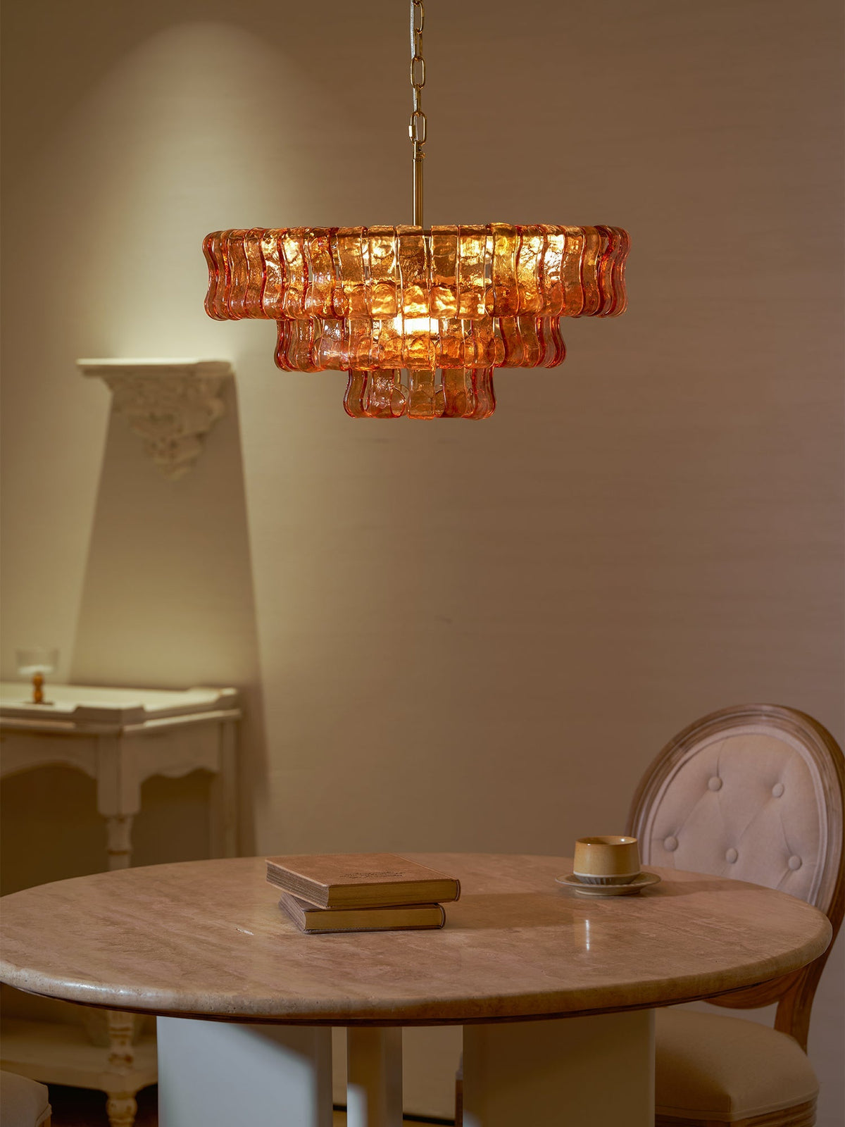 Warmly lit, this amber Clérachic chandelier hangs above a round table, with a cream chair back and side table in the shadows.