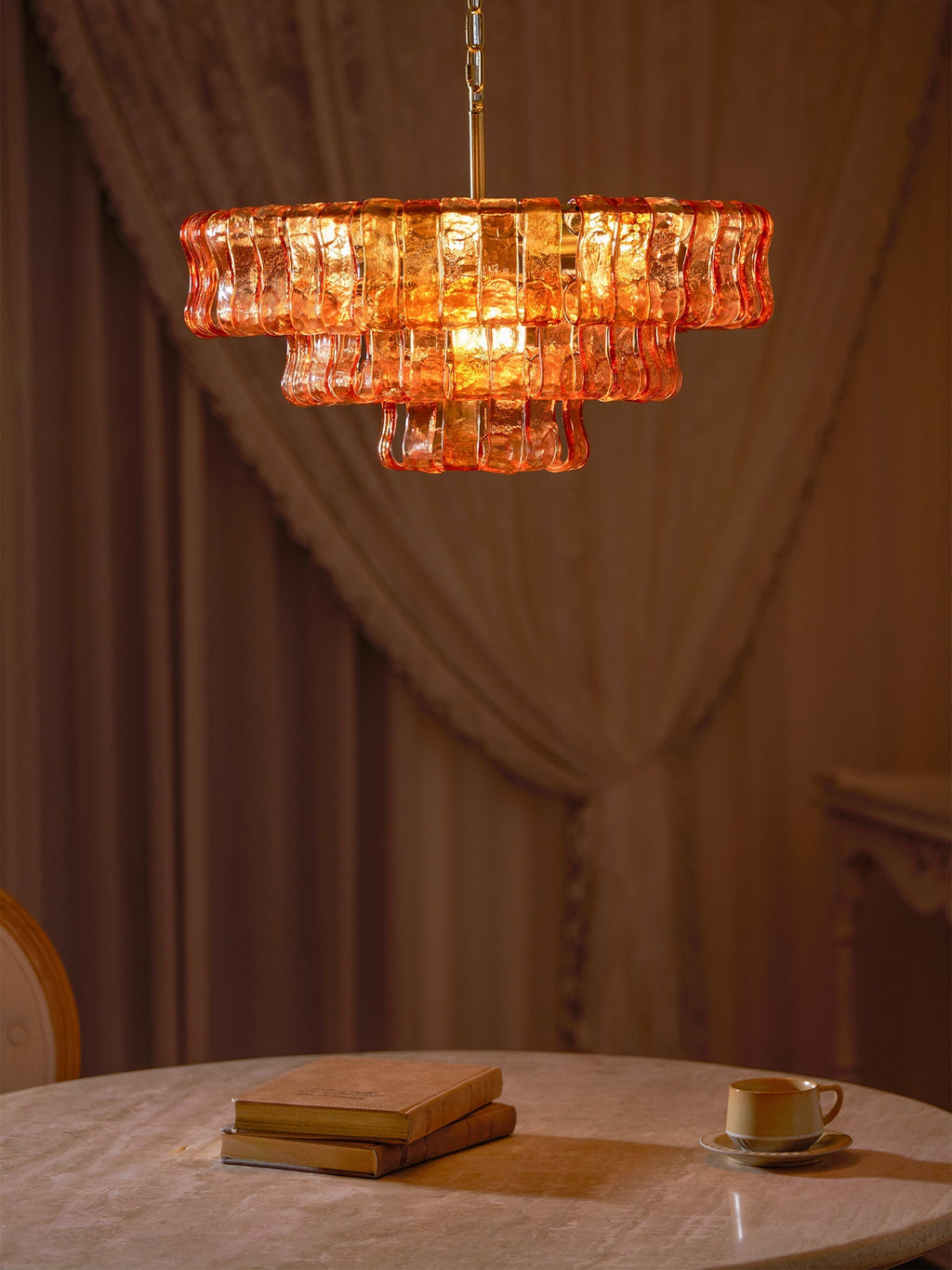 Brown velvet drapes frame this amber glass Clérachic chandelier, with a stack of books and a teacup on the table beneath.
