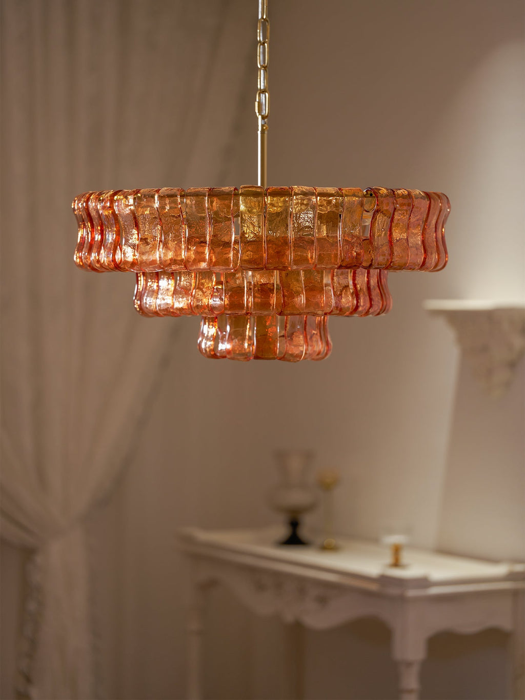 Seen from a distance, an amber glass chandelier from Clérachic hangs beside sheer curtains and a white console in a quiet room.