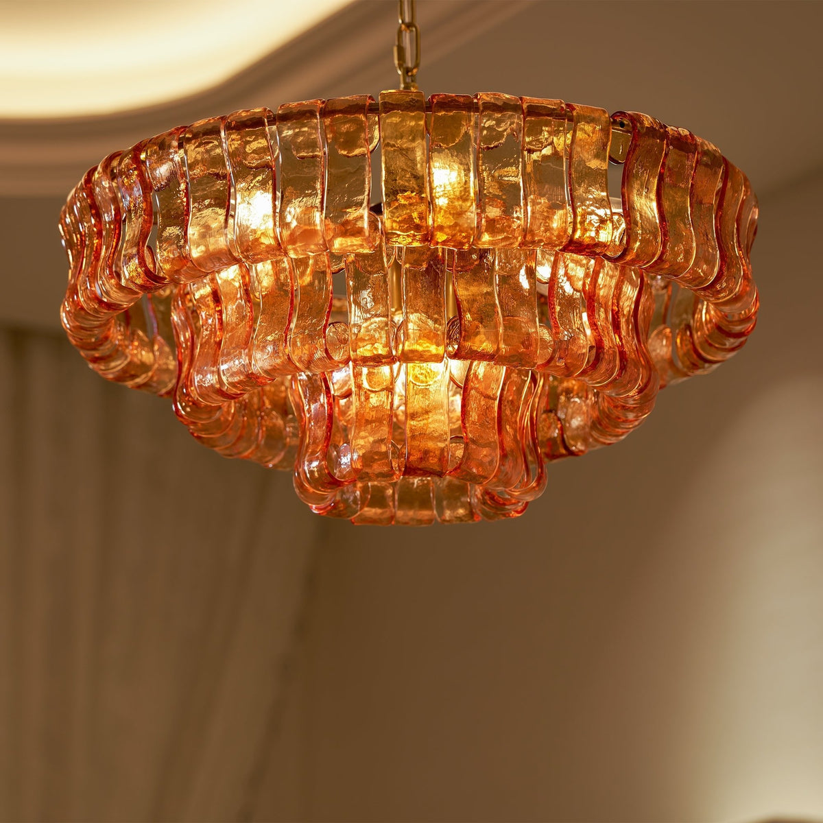 Looking up at an orange-pink glass Clérachic chandelier lit from within, its wavy glass tiles stacked in three rings.