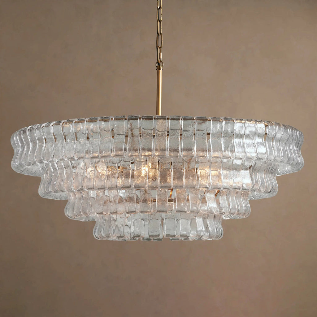 Clear textured glass tiers on a Clérachic chandelier with a brass stem, hung unlit against a warm taupe backdrop.