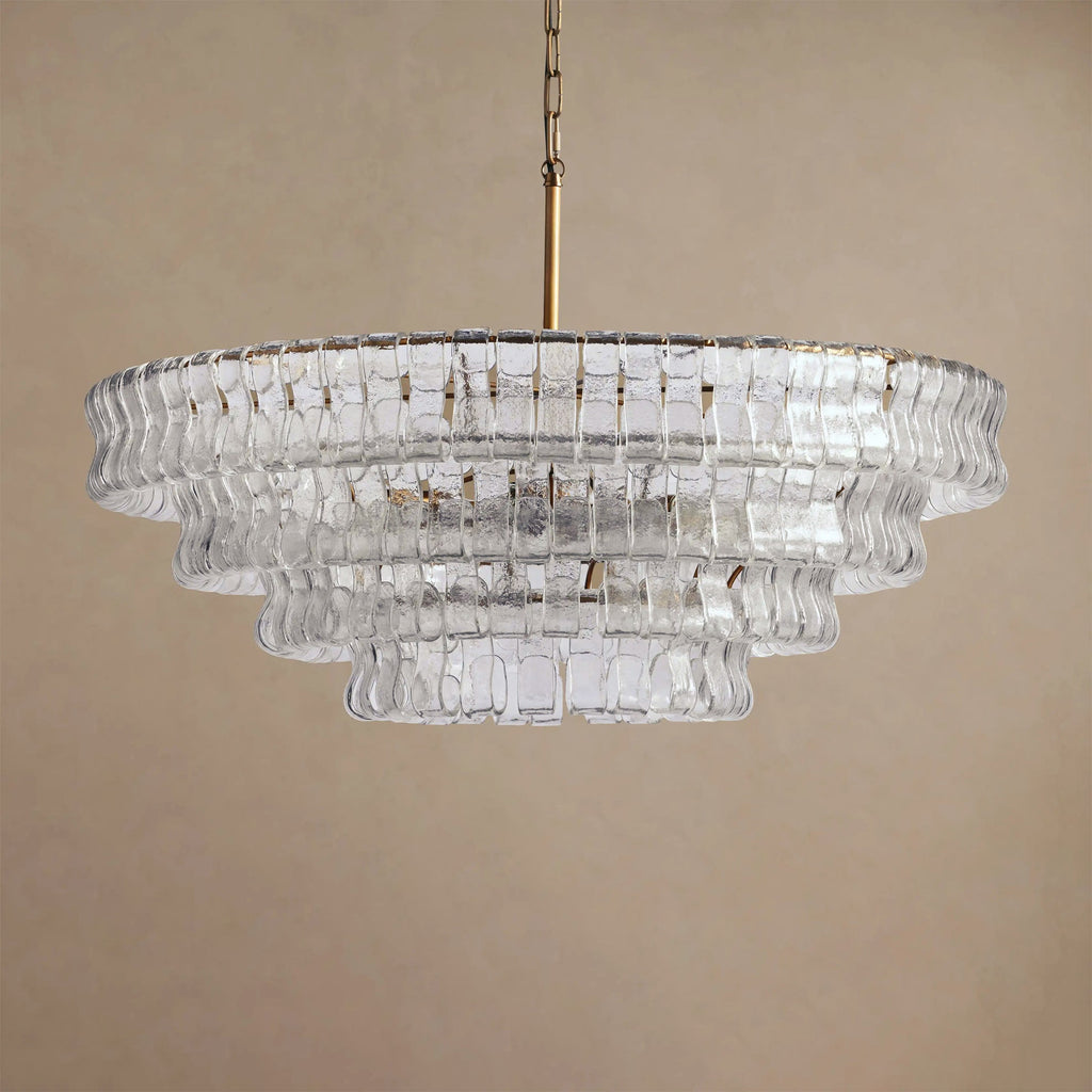 Clear glass chandelier from Clérachic, hung on a brass chain and stem in front of a sand-coloured backdrop.