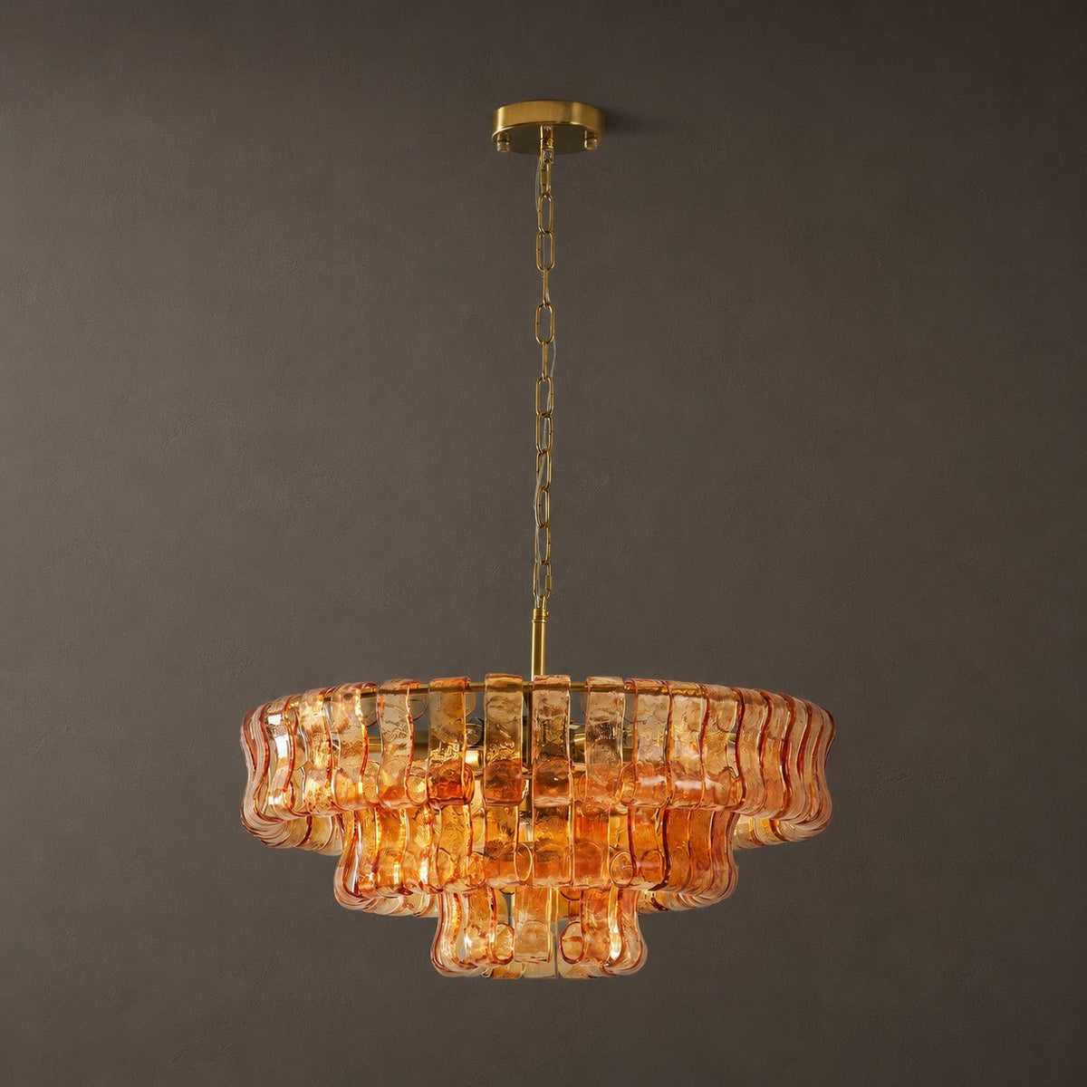 On a brass chain and round ceiling plate, this amber Clérachic chandelier is set against a dark grey wall.