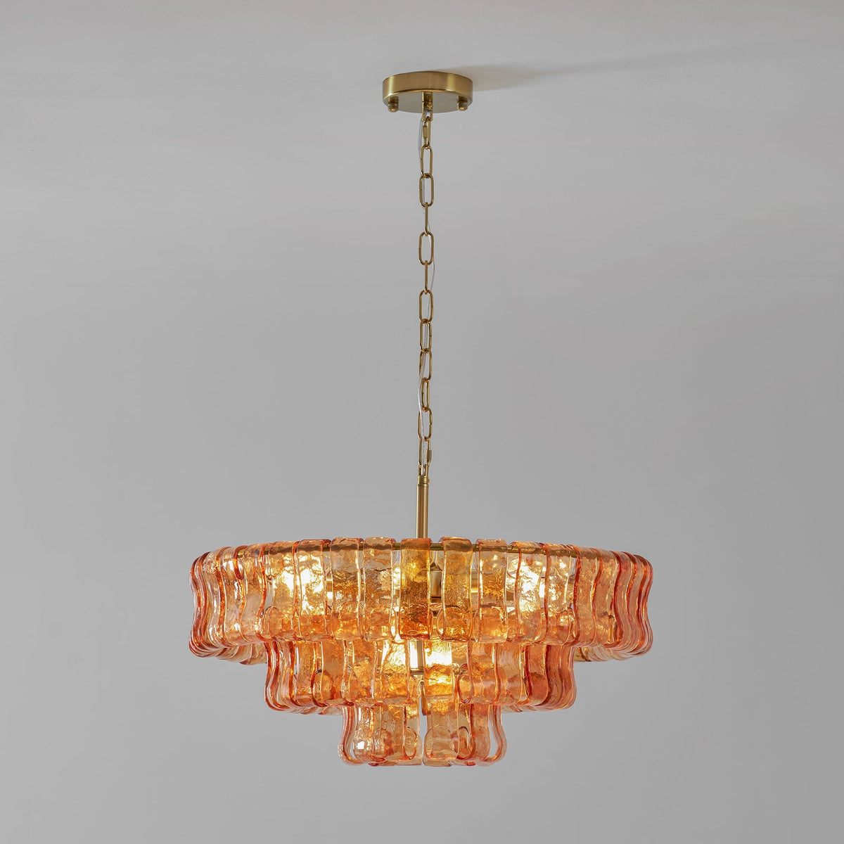 Amber-peach glass tiers on a Clérachic chandelier hung from a brass canopy and chain, against a light grey wall.