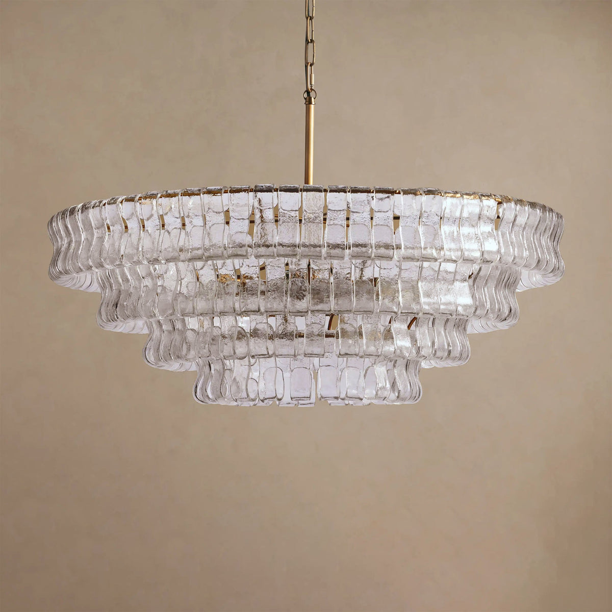 Pale clear glass tiers of a Clérachic chandelier with a brass stem, hung against a warm tan backdrop.