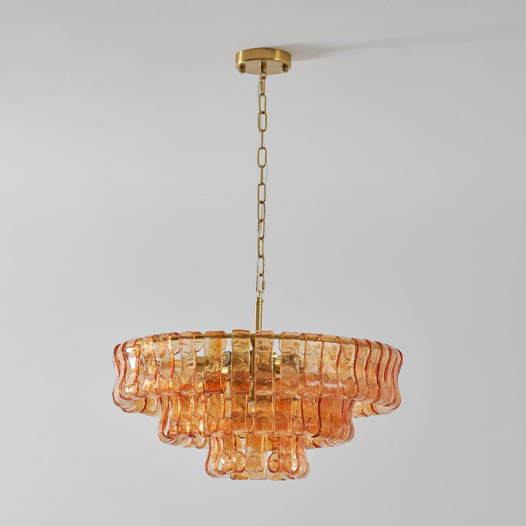 Peach-amber glass Clérachic chandelier hanging from a brass ceiling plate and chain, against a light grey wall.