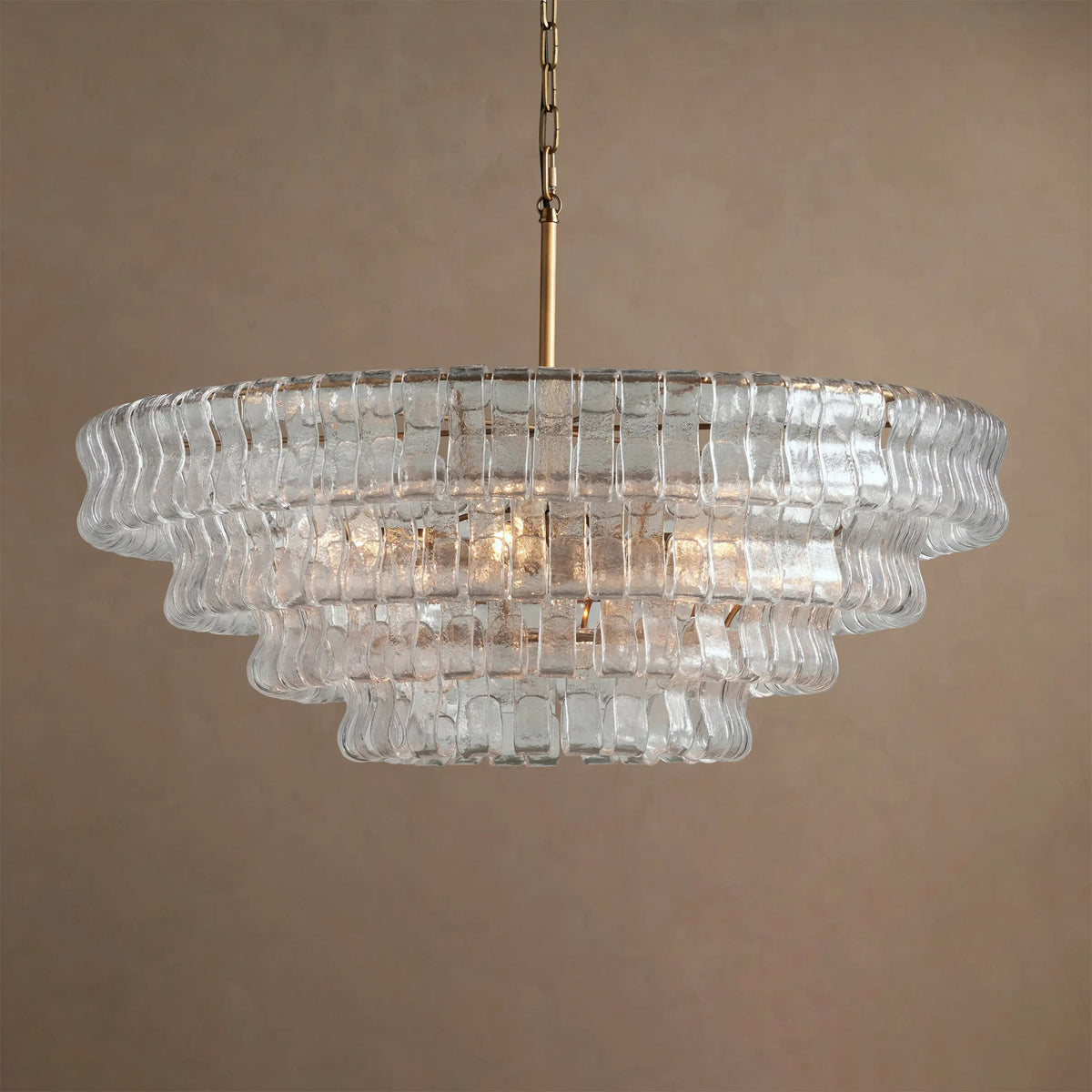Four tiers of clear glass on this Clérachic chandelier with a brass stem, hung against a warm beige backdrop.