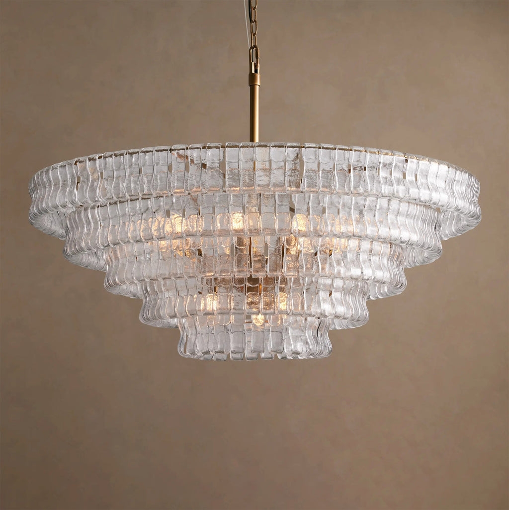 Five stepped tiers of clear glass with warm bulbs inside, on a Clérachic chandelier hung against a tan backdrop.