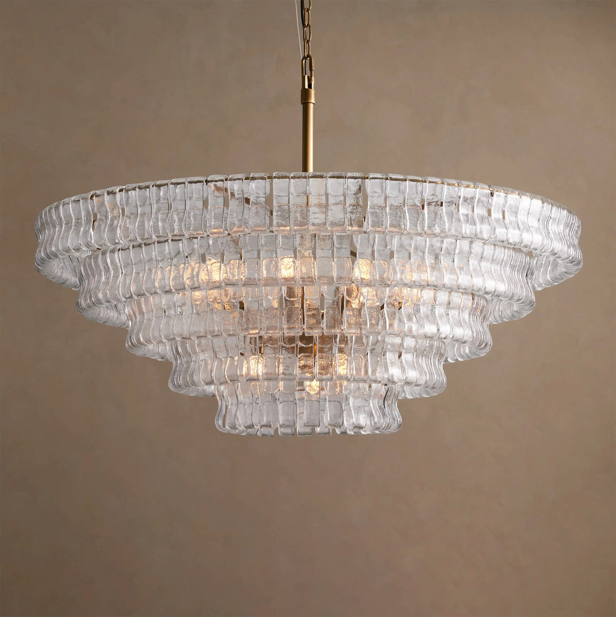 Five stepped tiers of clear glass with warm bulbs inside, on a Clérachic chandelier hung against a tan backdrop.