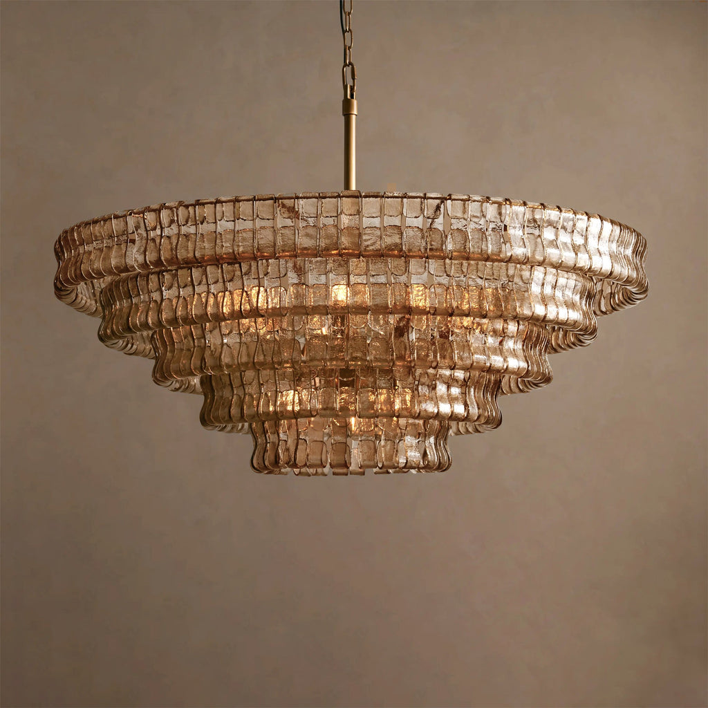 Champagne-toned glass tiers on a Clérachic chandelier with a brass stem, lit softly against a warm taupe backdrop.