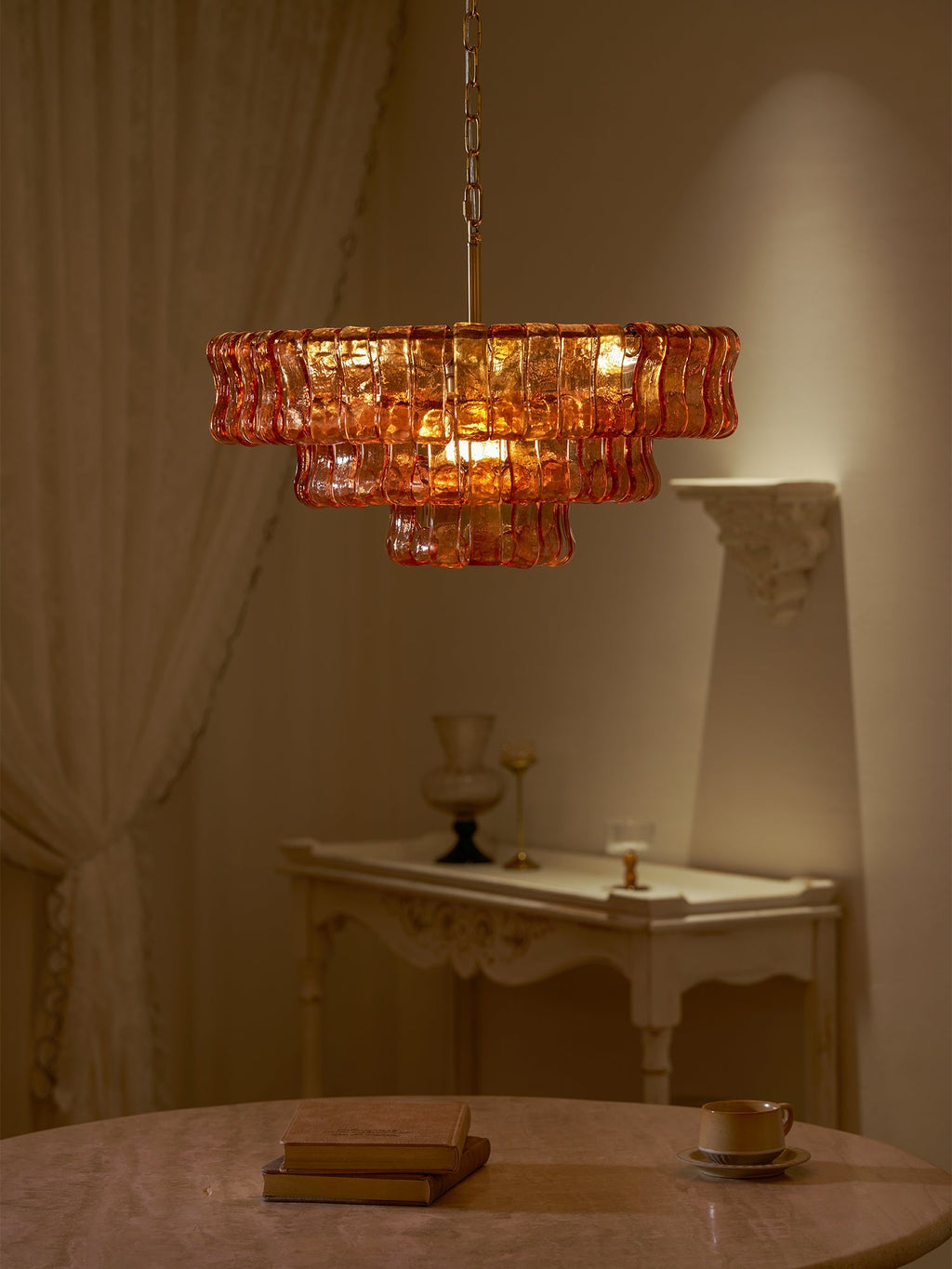 In a dim room, an amber Clérachic chandelier glows beside a white console and sheer curtain, with a book on the table.