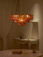 In a dim room, an amber Clérachic chandelier glows beside a white console and sheer curtain, with a book on the table.