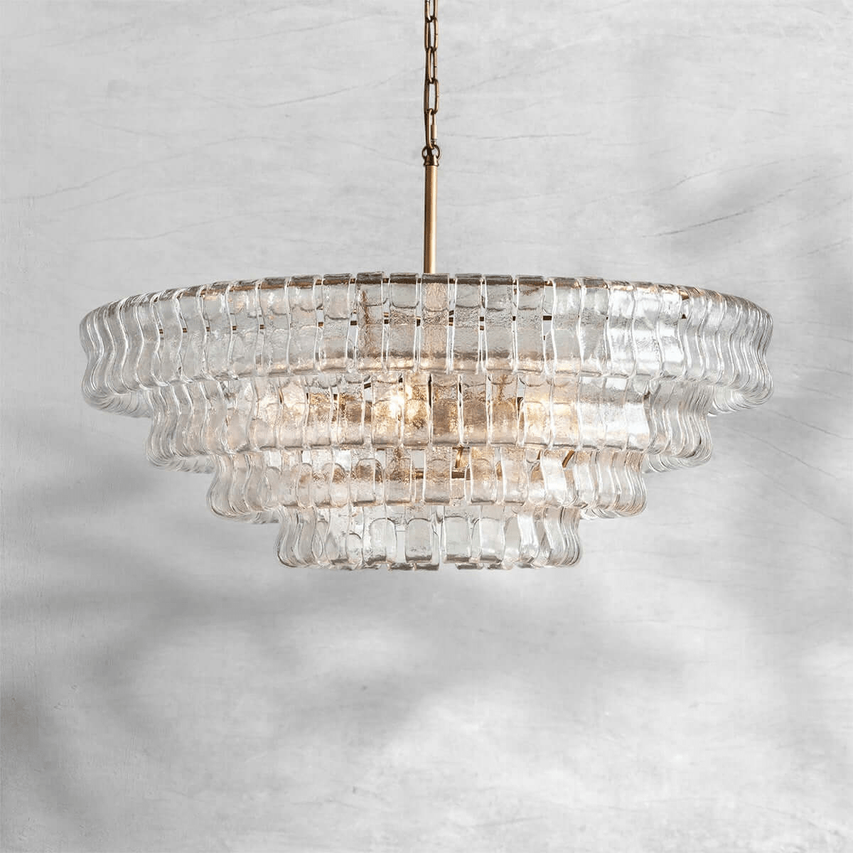 A clear glass Clérachic chandelier with a brass stem, shown against a pale marbled grey backdrop.