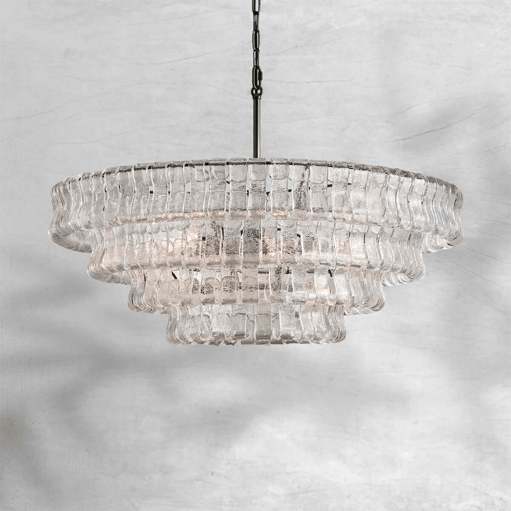 Wide and tiered, this clear-glass Clérachic chandelier hangs on a black chain against a pale marbled grey backdrop.