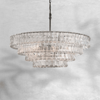 Wide and tiered, this clear-glass Clérachic chandelier hangs on a black chain against a pale marbled grey backdrop.