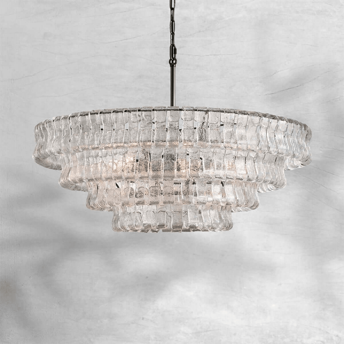 Wide and tiered, this clear-glass Clérachic chandelier hangs on a black chain against a pale marbled grey backdrop.