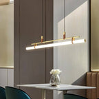 Clérachic gold linear pendant, tilted and lit, over a small white table with a glass vase of flowers in a grey-panelled room.