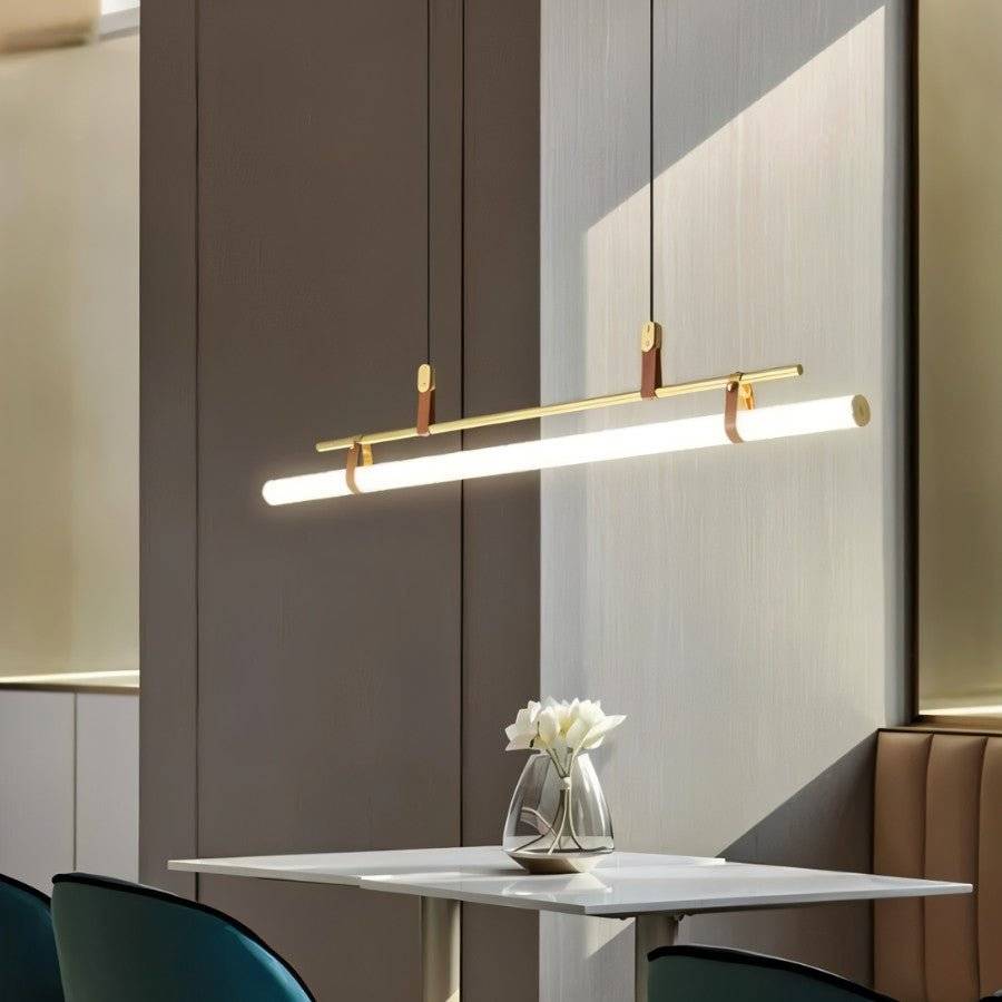 Clérachic gold linear pendant, tilted and lit, over a small white table with a glass vase of flowers in a grey-panelled room.