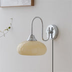 Clérachic glass wall lamp in chrome and beige, plug-in cord beside a wood frame and a trailing green vine.