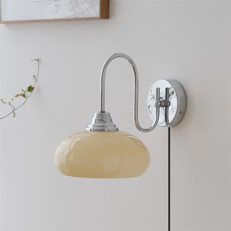 Clérachic glass wall lamp, chrome arm and beige glass, plug-in cord seen from a low angle beside a vine.