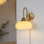 Clérachic glass wall lamp in copper with a lit beige shade and a plug-in cord, a plant soft in the background.