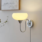 Clérachic glass wall lamp in chrome with a lit beige shade, plug-in cord beside a vine and a wood-framed print.