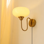 Clérachic glass wall lamp, copper arm and glowing beige glass, plug-in cord beside a soft curtain.