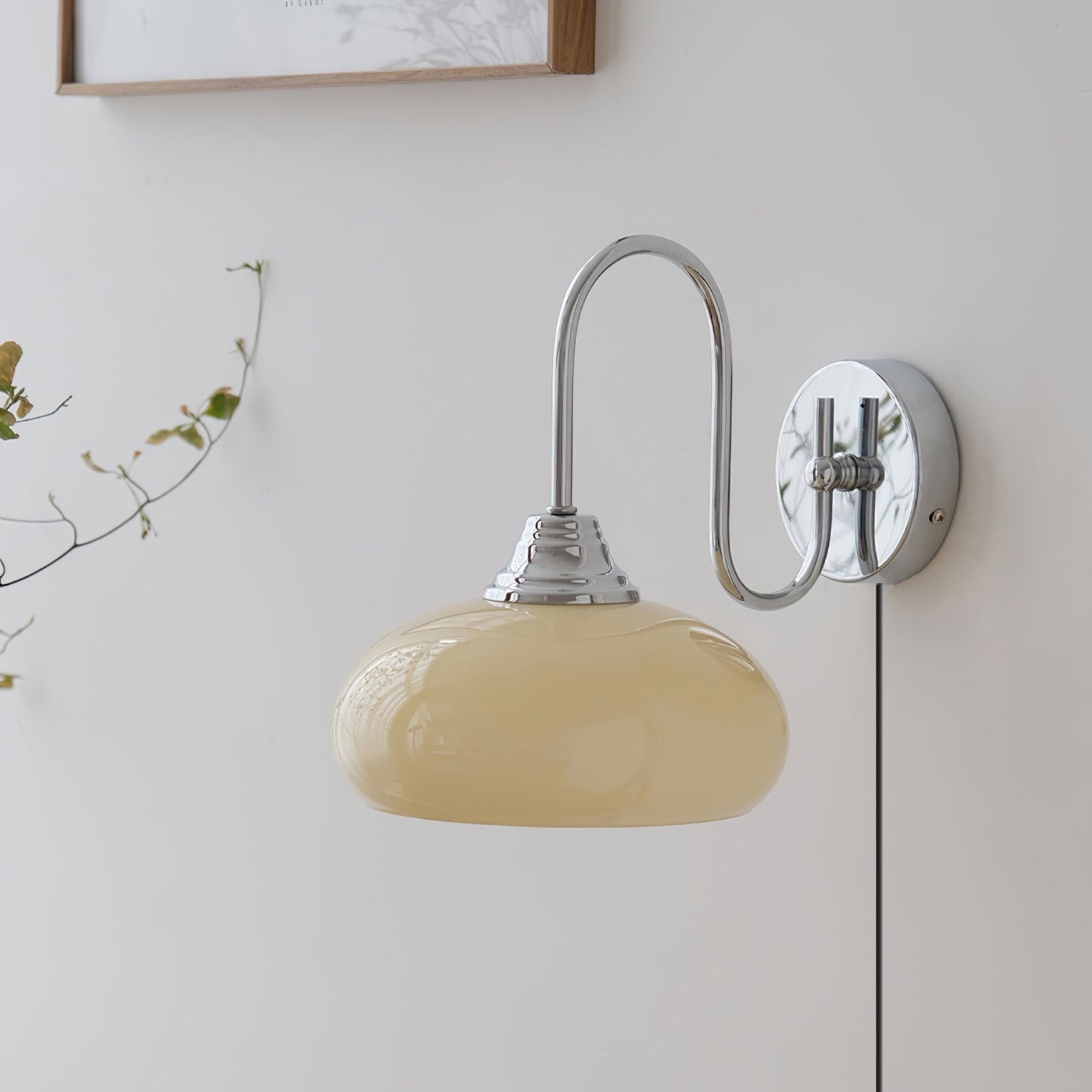 Clérachic glass wall lamp in chrome and beige, unlit plug-in version beside a wood frame and a leafy stem.