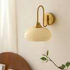 Clérachic glass wall lamp in copper and beige, hardwired unlit above a dark wood headboard by the curtain.