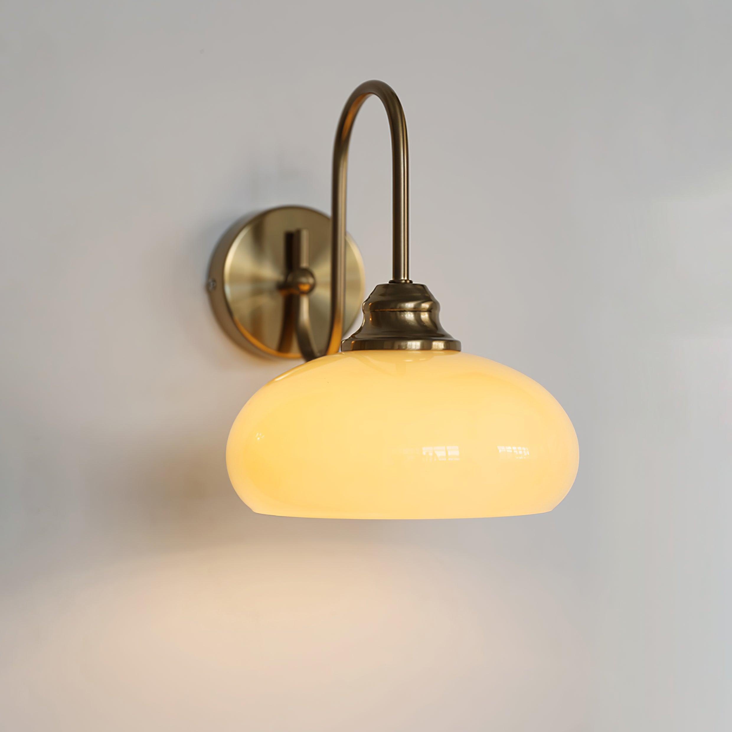 Clérachic glass wall lamp, copper J-arm and lit beige glass, hardwired with a bright pool under the shade.