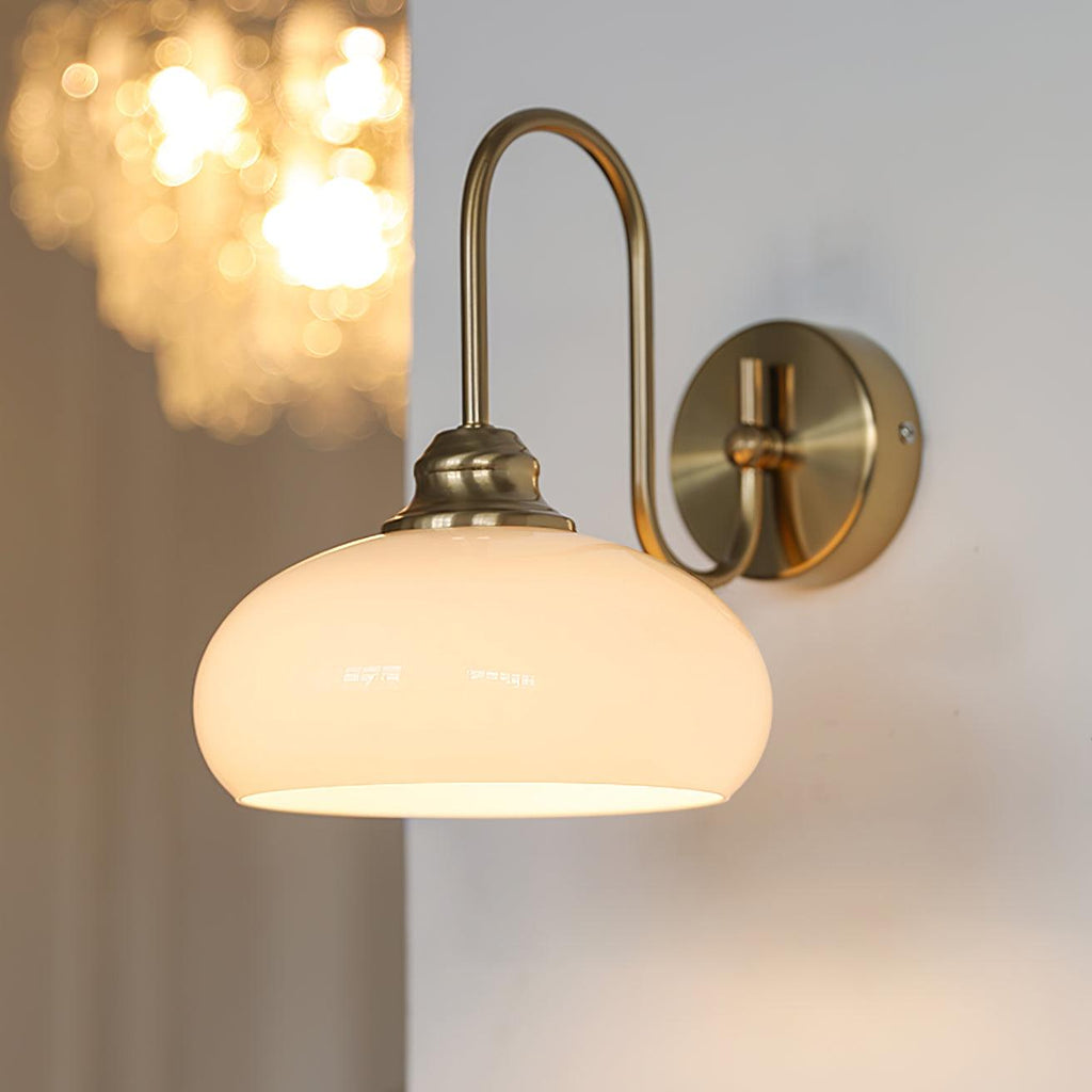 Clérachic glass wall lamp, copper crook arm and lit beige glass, hardwired with a warm light blurred behind.