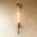 Clérachic wall light in aged gold with a slim fluted clear glass tube and a tall filament bulb glowing on a limewash wall.