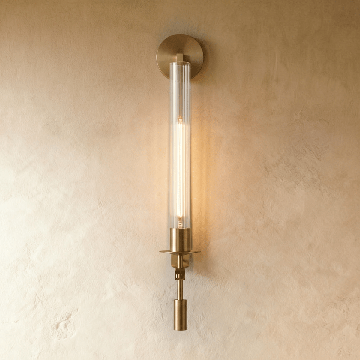 Clérachic wall light in aged gold with a slim fluted clear glass tube and a tall filament bulb glowing on a limewash wall.