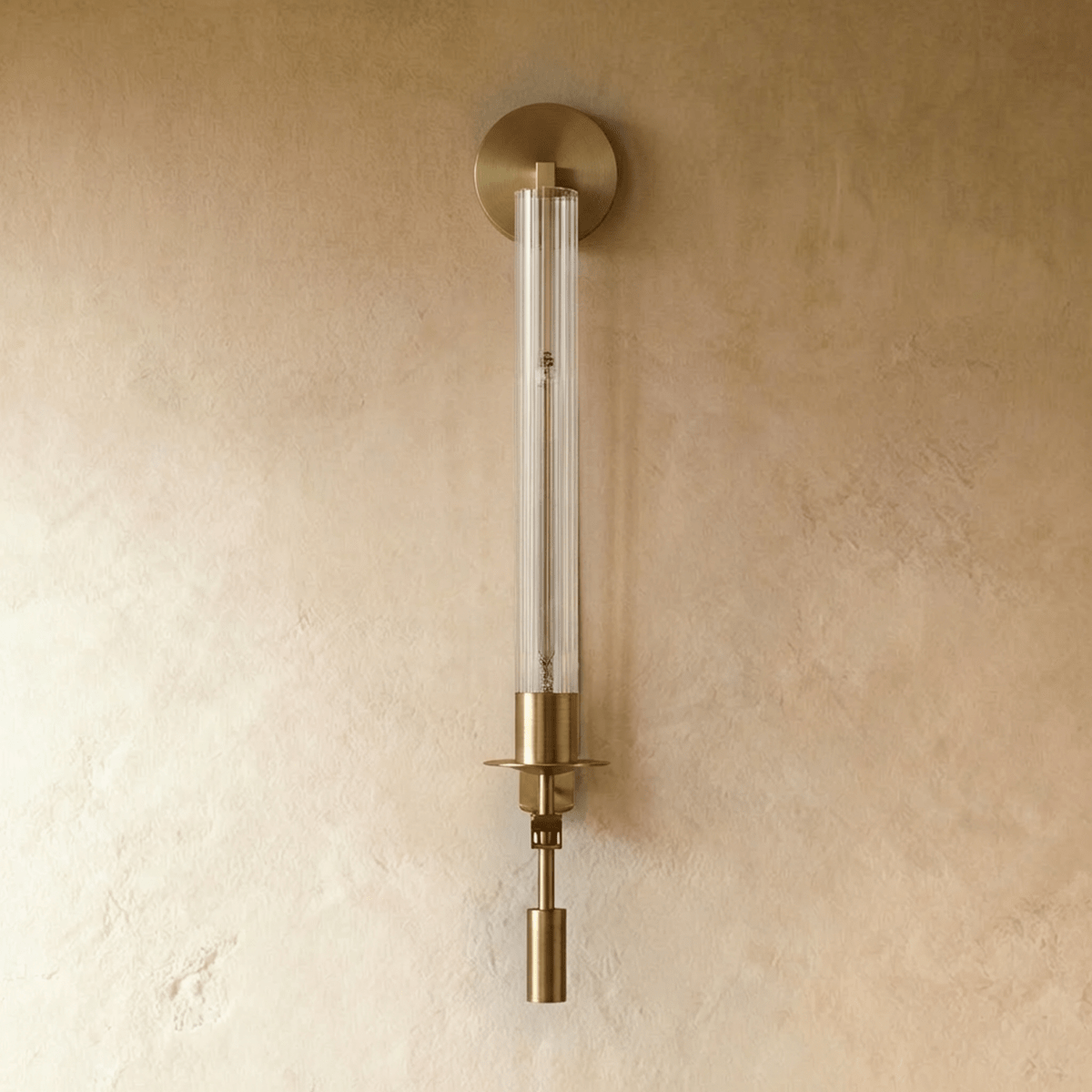 Clérachic wall light in aged gold with a fluted clear glass tube, round backplate and drop weight, shown unlit on limewash.